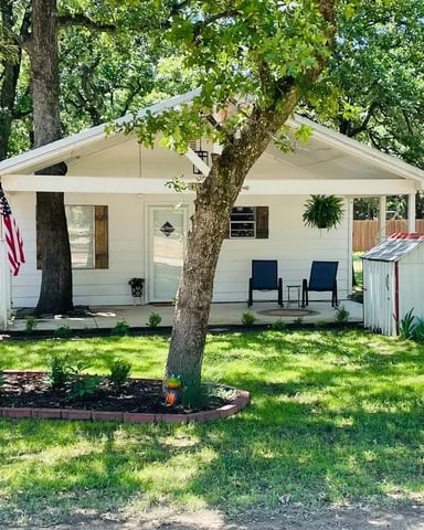 White cottage with covered porch, two blue chairs, large shade trees, and small shed in grassy yard