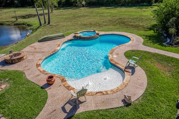 Backyard swimming pool with spa, concrete deck, lounge chairs, and fire pit overlooking a pond and lawn.