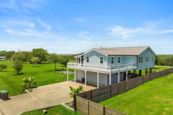 Light blue elevated house with white railings and covered porch on large green property with wooden fence