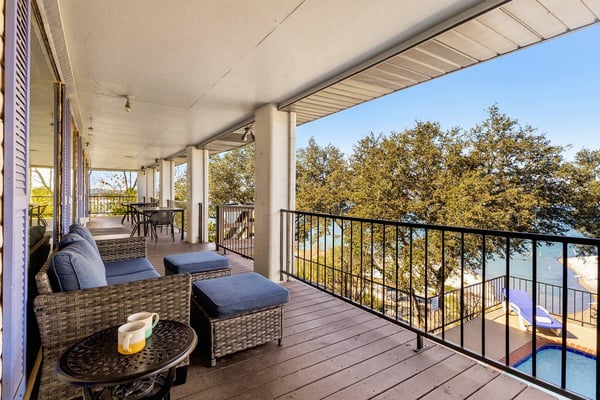Covered deck with wicker seating, lake view, and trees beyond black metal railing
