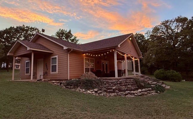 Single-story brown house with covered porch and string lights at dusk, stone fire pit in front yard, surrounded by trees.